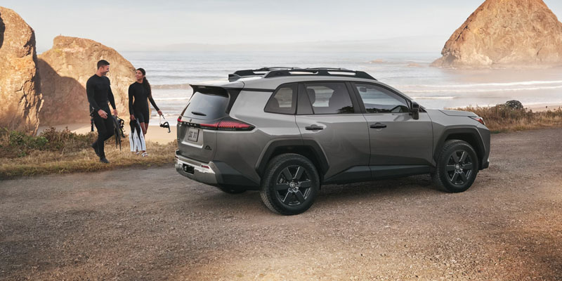 A grey SUV parked on a coastal road with two scuba divers walking toward the beach